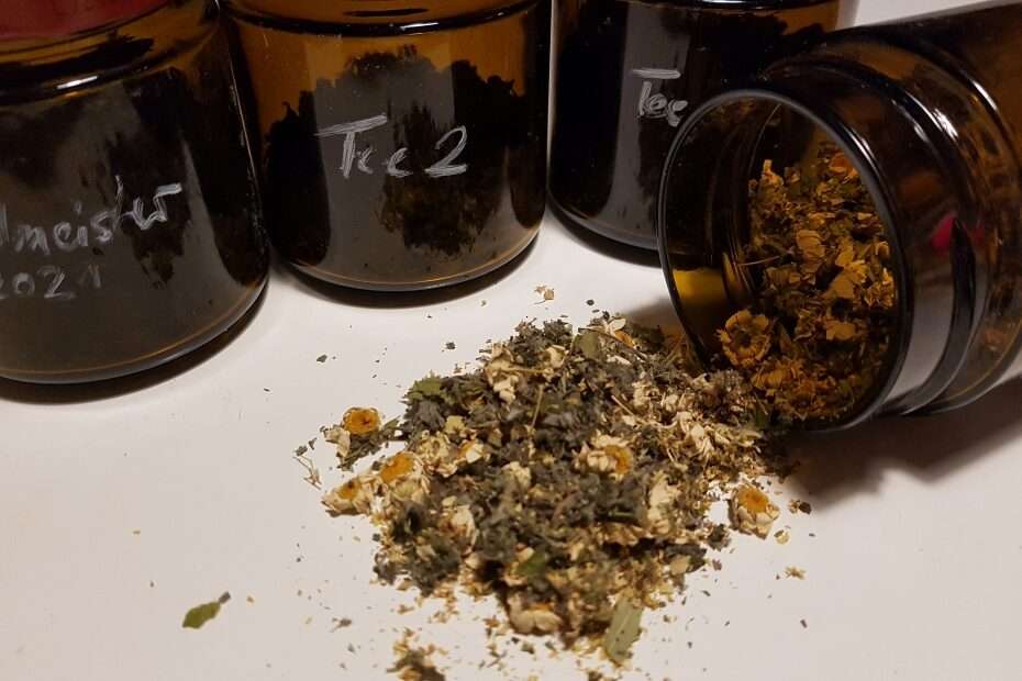 4 braune Gläser mit selbstgemachten Teemischungen das rechte Glas ist offen und liegt aus der Seite, Teile der Teemischung liegen auf dem Tisch. Man sieht getrocknete Kamillenblüten und kleine getrocknete Blätterteile