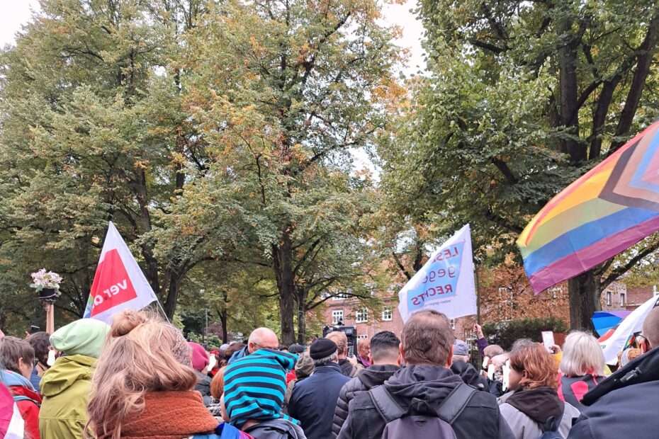 Menschen bei der Demo gengen die todestrafe für zwei Lesben im Iran von hinten. Eine LGBTQIA+ Fahre und eine Queer VERDI Fahne sind zu sehen.