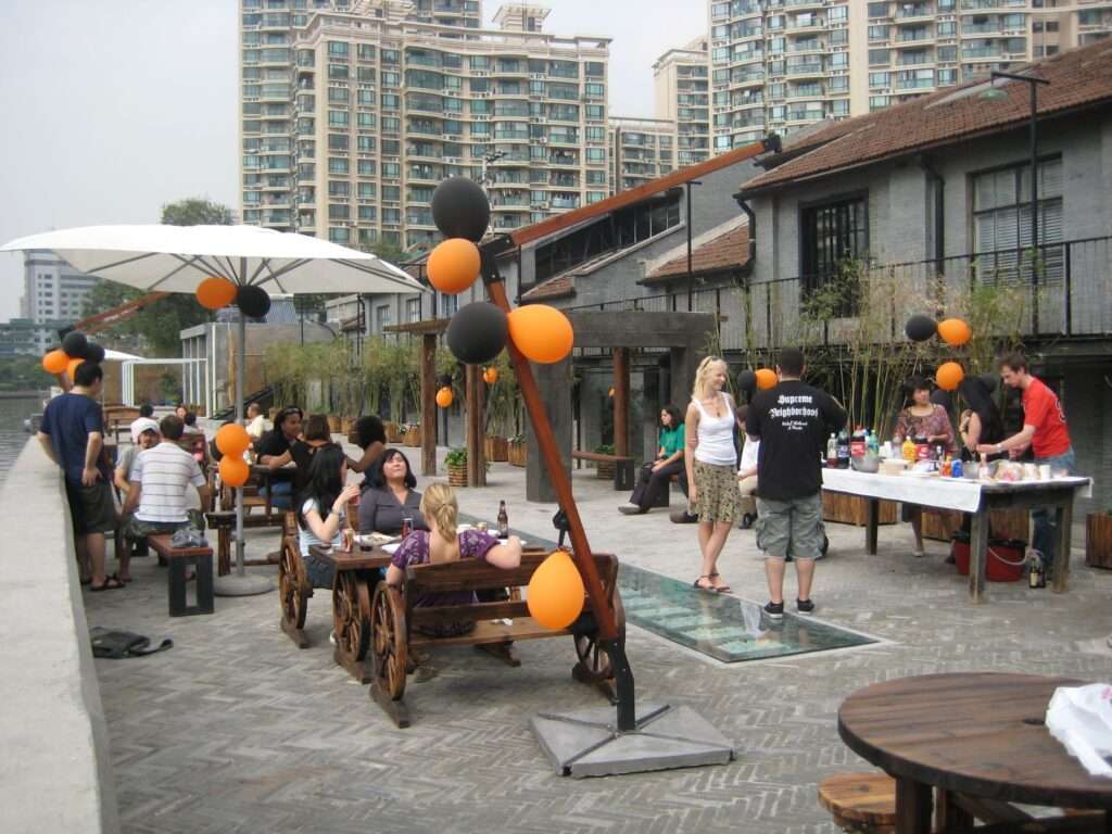 A terrace next to the river where people sit on wooden benches. There is a white parasol and orange and black baloons as decoration. On the right there is a table with lots of drinks and bottles. In the background are big apartment buildings.