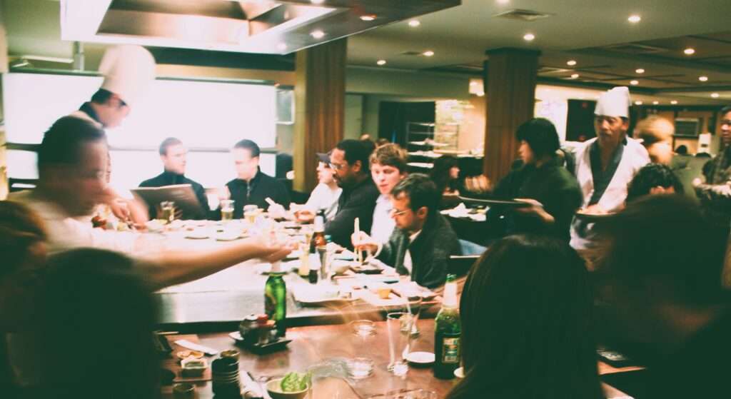 The picture is blurry, it shows a restaurant. People are sitting around a square table with a teppanyaki cooking plate in the middle and a chef. Servcers are bringing more food.