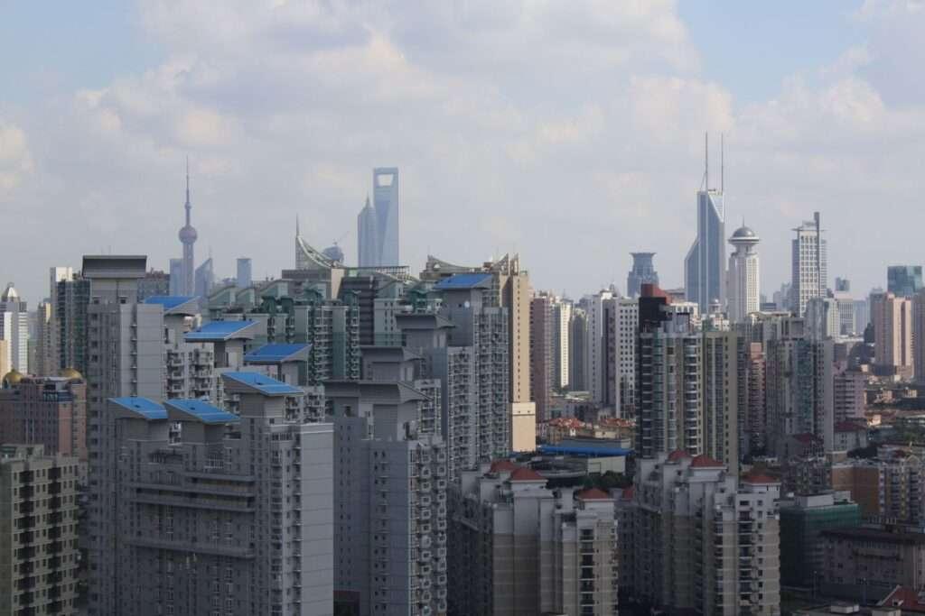 Many mostly grey apartment buildings with around 20 floors or more. There are patches of blue sky and white clowds in the background a few iconic building of the Shanghai Skyline are visible - like the Oriental Pearl Tower.