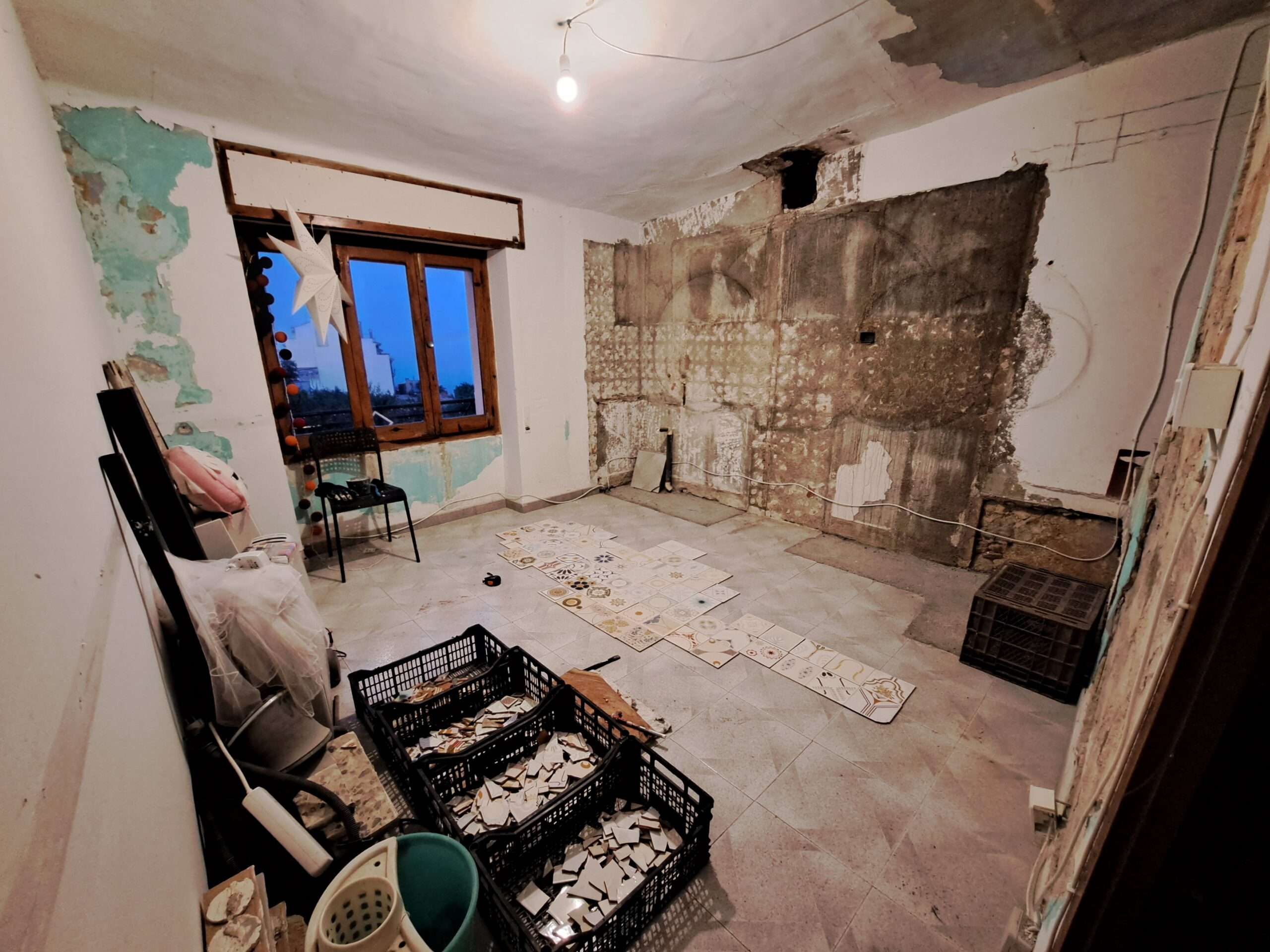 Wide angel view of the room. There are boxes with tile pieces, a window. The walls are patchy and colour and plaster is coming off everywhere.
